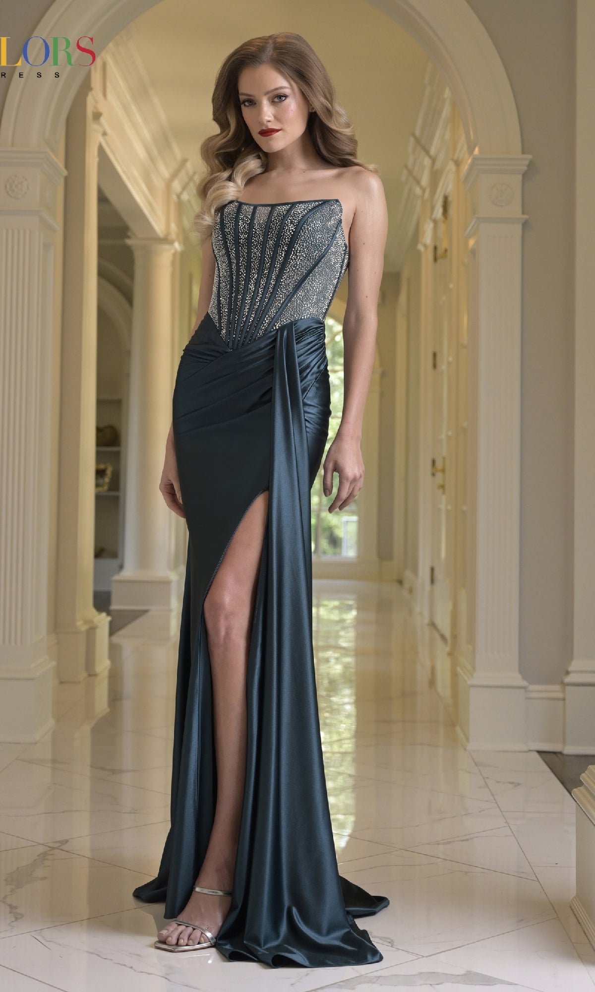 A woman wears the Strapless Long Corset Prom Gown by Colors Dress (style 3933), featuring a fitted, dark teal embellished bodice, as she stands in an elegant hallway with marble floors and classical columns.